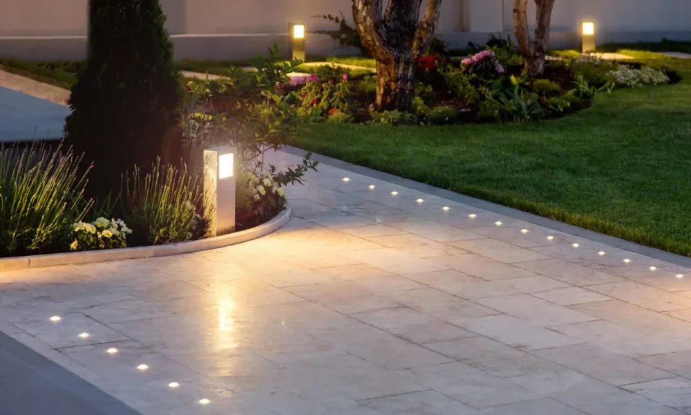 A modern outdoor garden pathway featuring sleek tile flooring with embedded LED lights for illumination. The path is bordered by well-maintained greenery and subtle automated bollard lights, creating an elegant and functional lighting solution.